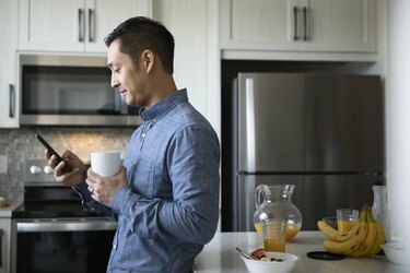 Mies juo kahvia ja käyttää älypuhelinta aamukeittiössä