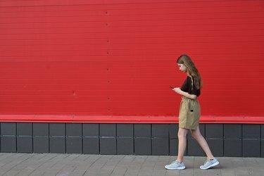 Портрет молодой студентки, читающей сообщение по мобильному телефону, стильной красивой девушки, болтающей по мобильному телефону во время прогулки по улице