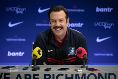 Тед Лассо сидить за столом для прес-конференції в 3 сезоні.