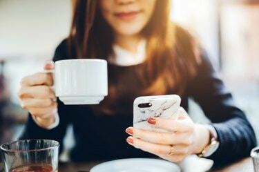 Iš arti jaunos moters, geriančios kavą ir skaitančios naujienas mobiliajame telefone anksti ryte prieš darbą
