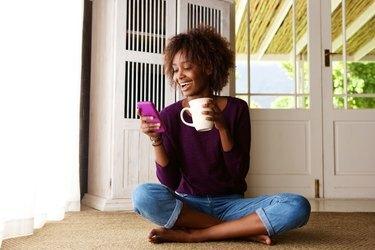 Besišypsanti moteris sėdi ant grindų namuose su mobiliuoju telefonu