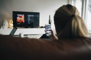 Vista trasera de una adolescente usando la aplicación de teléfono inteligente mientras ve la televisión en la sala de estar en casa