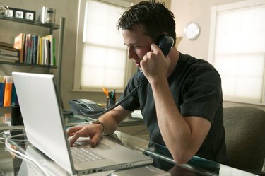 um jovem caucasiano sentado trabalhando em seu computador e ao telefone em seu escritório em casa