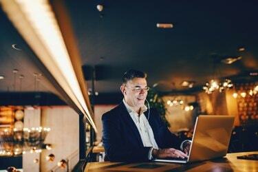 Om de afaceri de vârstă mijlocie zâmbitor folosind laptop și având căști în urechi în timp ce stă seara în cafenea.