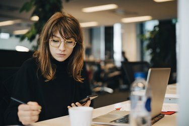 Forretningskvinne skriver mens hun holder mobiltelefonen ved skrivebordet på kontoret