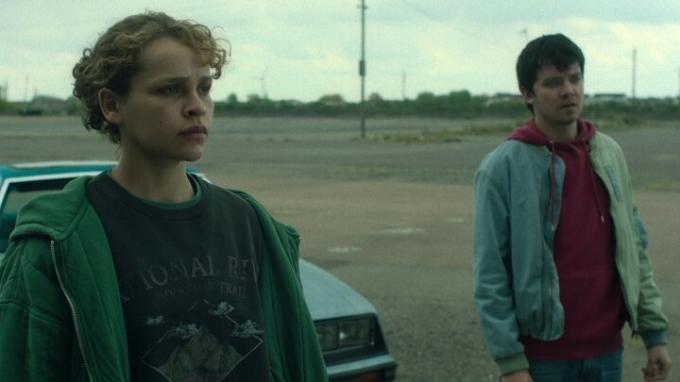 Iola Evens und Asa Butterfield stehen in „Choose Or Die“ vor einem Auto auf einem leeren Parkplatz.