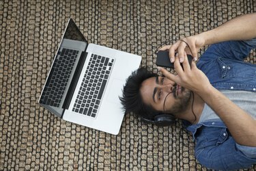 Azijos vyras guli ant grindų su nešiojamu kompiuteriu ir telefonu.