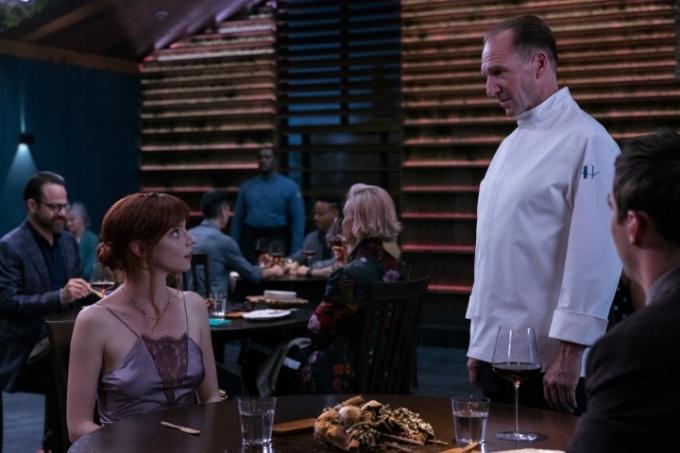 Anya Taylor-Joy gaat zitten en kijkt op naar Ralph Fiennes in een scène uit The Menu.