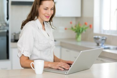Leende ung kvinna som använder bärbar dator i köket