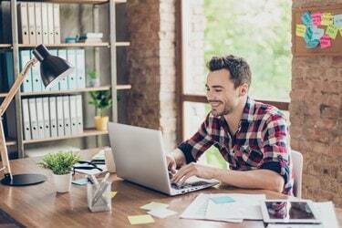 Alegre joven brunet freelancer está sonriendo, escribiendo en su computadora portátil en una agradable y moderna estación de trabajo en casa, en ropa casual elegante