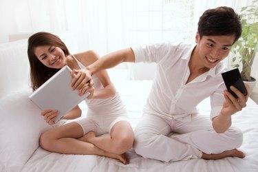 Млади пар који гледа мобилни телефон и таблет рачунар у кревету