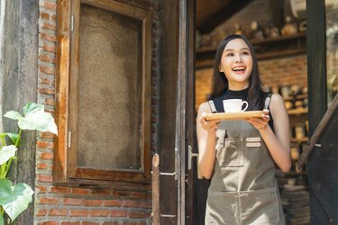 カフェの女性オーナーは、顧客のために開く準備ができている肯定的な幸福に微笑み、コーヒーショップの入り口に立っているエプロンハンドプレゼントトレイとコーヒーカップの温かい歓迎の自信を持って自信を持って立っているアジアの女性