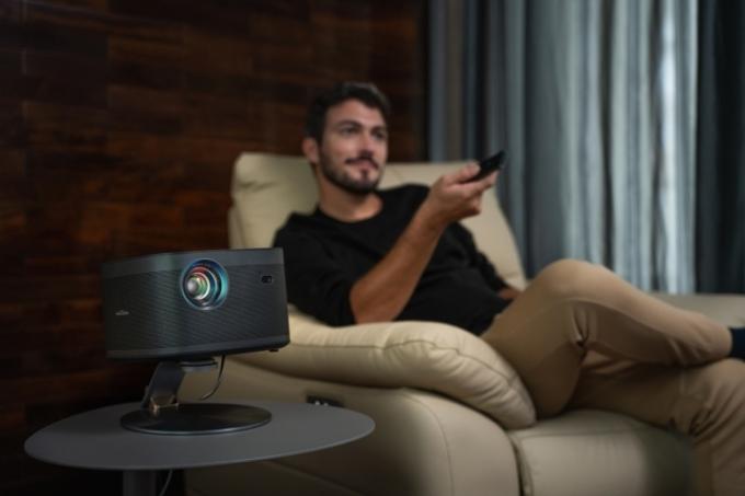 Un hombre en una silla con un control remoto y un proyector sobre una mesa. 