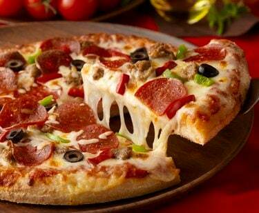 شريحة بيتزا يتم رفعها من فطيرة بيتزا بها الكثير من خيوط الجبن والمكونات في الخلفية.