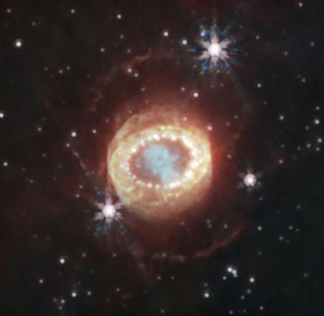 Камера NIRCam (ближняя инфракрасная камера) Уэбба сделала это детальное изображение SN 1987A (Сверхновая 1987A). В центре материал, выброшенный сверхновой, образует форму замочной скважины. Слева и справа от него видны слабые полумесяцы, недавно обнаруженные Уэббом. За ними экваториальное кольцо, образовавшееся из материала, выброшенного за десятки тысяч лет до взрыва сверхновой, содержит яркие горячие точки. За пределами этого находится диффузное излучение и два слабых внешних кольца. На этом изображении синий представляет свет длиной 1,5 микрона (F150W), голубой — 1,64 и 2,0 микрона (F164N, F200W), желтый — 3,23 микрона (F323N), оранжевый — 4,05 микрона (F405N) и красный — 4,44 микрона (F444W).