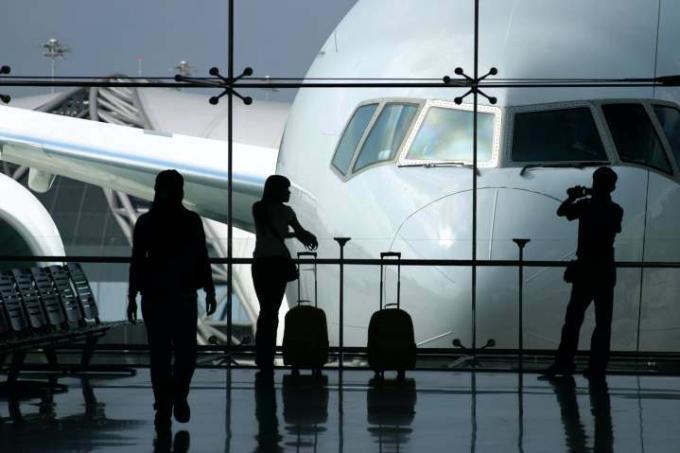 Τρεις σκιαγραφημένοι επιβάτες στον χώρο αναμονής του αεροδρομίου με μεγάλο αεροσκάφος στο φόντο.
