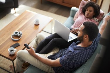 Pareja sentada en un sofá en casa con ordenador portátil y viendo la televisión