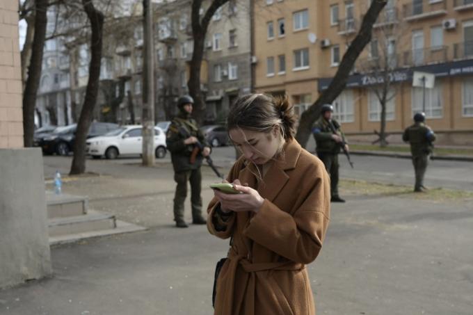 Eine Frau blickt auf ihr Mobiltelefon in der Nähe von Soldaten, die am 29. März 2022 in Mykolajiw, während der russischen Invasion in der Ukraine, vor einem Regierungsgebäude Wache halten, das von russischen Raketen getroffen wurde.