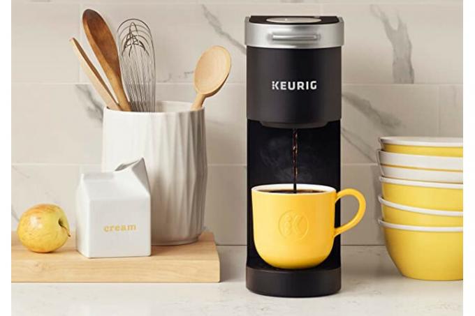 Кофеварка Keurig K-Mini варит кофе в желтой кружке, стоящей на кухонном столе рядом с кадкой с кухонной утварью.