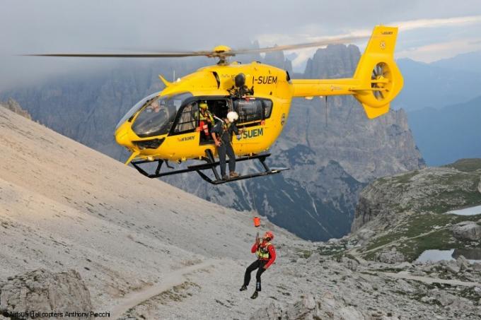 Um homem está pendurado em um cabo de um helicóptero amarelo, com montanhas ao fundo.