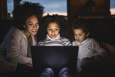 घर में सोफे पर लैपटॉप का उपयोग करते हुए मुस्कुराते हुए अफ्रीकी अमेरिकी मां और उनके बच्चे।