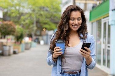 Mujer feliz bebiendo una taza de café en movimiento mientras envía mensajes de texto en su teléfono