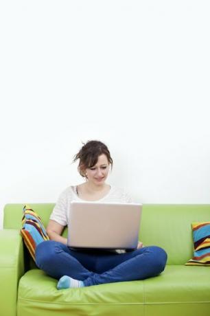 Пълна дължина на щастлива млада жена, използваща лаптоп, докато седи на зелен диван