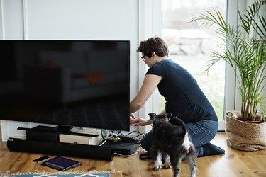 Беременная женщина прокладывает кабели телевизора, стоя на коленях у собаки в гостиной