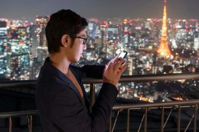 Άνθρωπος που χρησιμοποιεί smartphone ενώ στέκεται στο μπαλκόνι με θέα στον ορίζοντα της πόλης τη νύχτα.