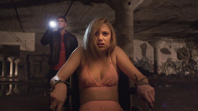 Maika Monroe przywiązana do krzesła, a za nią mężczyzna świecący latarką w It Follows.