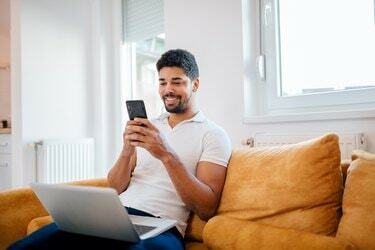 Насмејани слободњак мешане расе који користи паметни телефон и лаптоп док седи на софи.