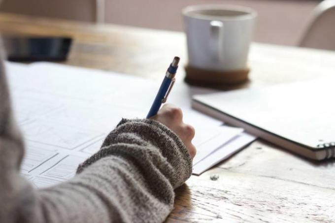 Una persona che studia e scrive appunti su un pezzo di carta. C'è una tazza di caffè e un raccoglitore in lontananza.