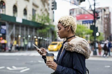 JAV, Niujorkas, madinga jauna moteris su kava eina žiūrėti į mobilųjį telefoną gatvėje