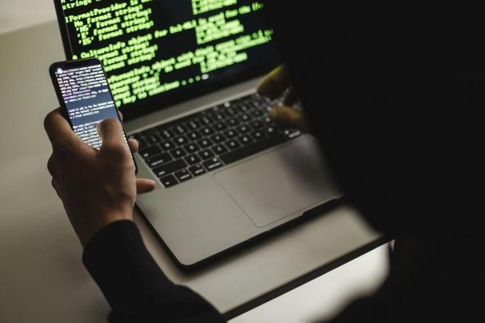 Хакер друкує на ноутбуці Apple MacBook, тримаючи телефон. Обидва пристрої показують код на своїх екранах.
