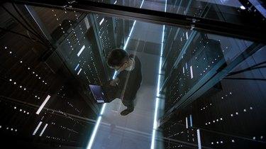 Pohled shora přes sklo IT inženýra pracujícího s notebookem v datovém centru plném aktivních rackových serverů.