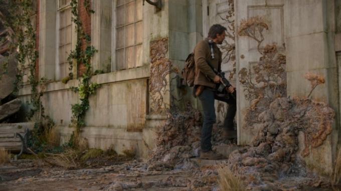 Joel segura um rifle à sua frente ao entrar por uma porta coberta de fungos secos em The Last of Us na HBO. 