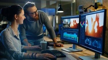 Vídeo feminino e editor de som trabalha com seu colega masculino em um projeto em seu computador pessoal com dois monitores. Eles trabalham em um escritório criativo Loft.