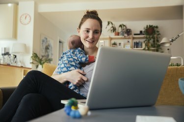 노트북을 사용하고 집에서 갓난 아기를 안고 있는 어머니