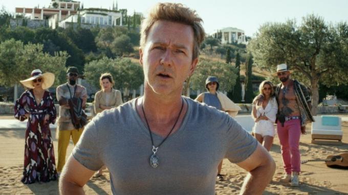 『ナイブズ アウト 2』でエドワード ノートンがビーチに立っており、後ろに人々がいます。