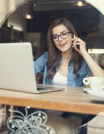 Kvinne snakker med mobiltelefon og bruker bærbar PC på kafé
