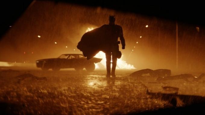 Batman nadert de camera in een regenachtige straat met een vuur op de achtergrond.