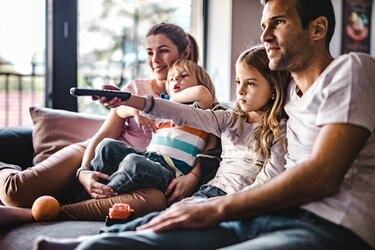 Jauni tėvai mėgaujasi savo mažais vaikais ant sofos kartu žiūrėdami televizorių.