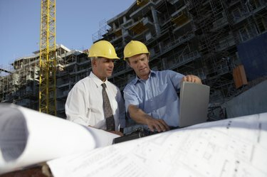 Dva muži v přilbě dívají se dolů na přenosný počítač na staveništi