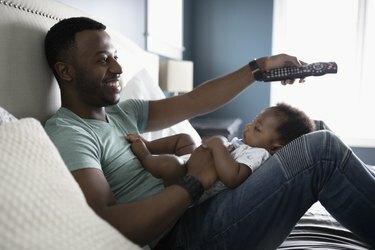 يبتسم الأب مع جهاز التحكم عن بعد يشاهد التلفزيون مع ابنه الرضيع في حضنه على السرير