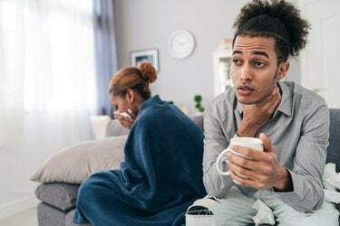 病気の若いアフリカ系アメリカ人のカップルが自宅のソファに座って、頭痛の種の毛布で覆われている、男性が温かい飲み物でカップを持って喉が急上昇しているショット