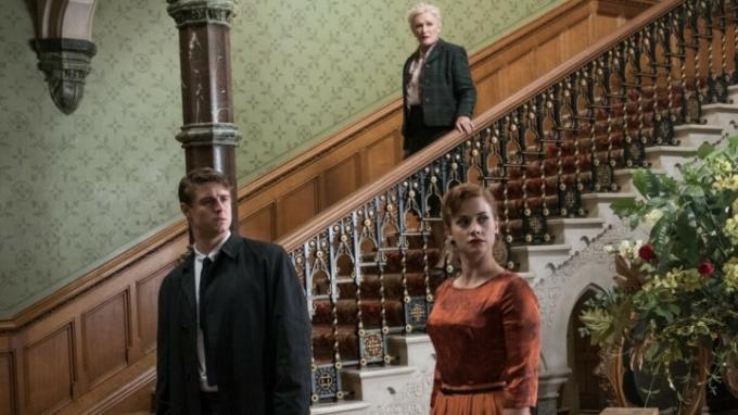 Una mujer baja una escalera mientras dos personas esperan abajo en Crooked House.