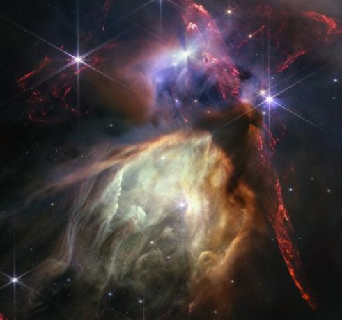 Het eerste jubileumbeeld van de NASAESACSA James Webb Space Telescope toont de geboorte van een ster zoals het nog nooit eerder is gezien, vol gedetailleerde, impressionistische textuur. Het onderwerp is het Rho Ophiuchi-wolkencomplex, het dichtst bij de aarde gelegen stervormingsgebied. Het is een relatief kleine, stille stellaire kinderkamer, maar je zou het nooit weten uit de chaotische close-up van Webb. Jets die uit jonge sterren barsten, doorkruisen het beeld, botsen op het omringende interstellaire gas en verlichten moleculaire waterstof, weergegeven in rood. Sommige sterren vertonen de veelbetekenende schaduw van een circumstellaire schijf, de ingrediënten van toekomstige planetaire systemen.