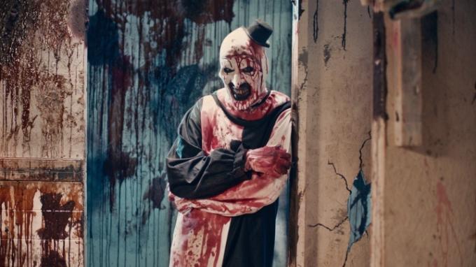 يتكئ Art the Clown على الحائط ويحدق بشكل مخيف في لقطة ثابتة من فيلم Terrifier 2.