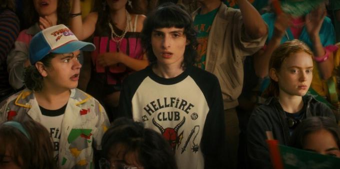 Gaten Matarazzo, Finn Wolfhard y Sadie Sink se paran entre la multitud durante un mitin de la escuela secundaria en una escena de Stranger Things 4.