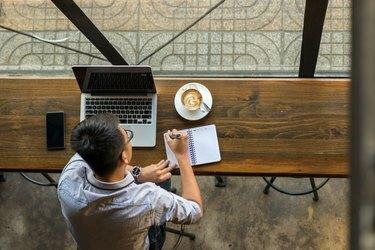 Молодой азиатский бизнесмен, принимая к сведению рядом с ноутбуком на деревянном столе